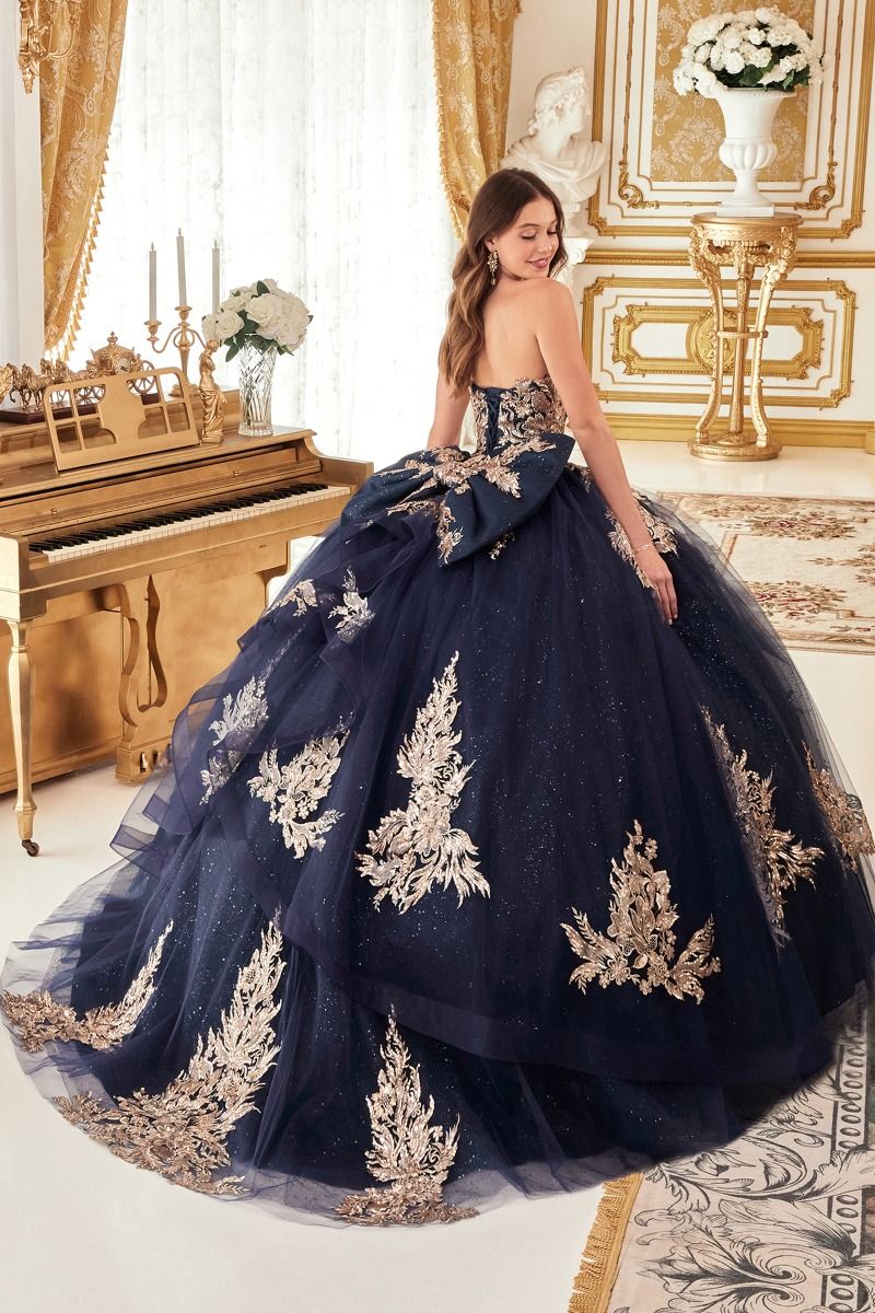 A woman in the Ladivine Strapless Quinceanera Dress 15715 with gold embroidery stands beside a gold piano in an elegant white and gold room.